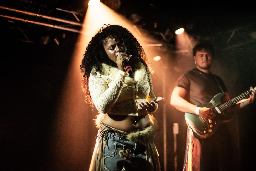 A female singer with curly hair performs passionately on stage, holding a microphone, while a male guitarist plays in the background under dramatic lighting.