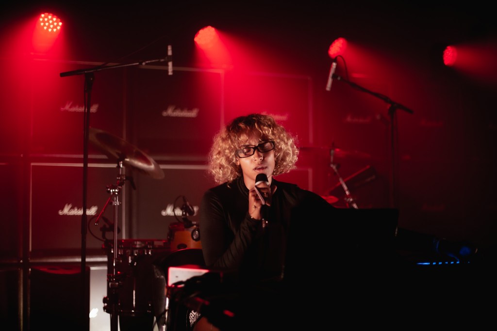 A musician performing on stage with a microphone, surrounded by musical instruments and amplifiers, lit by red stage lights.