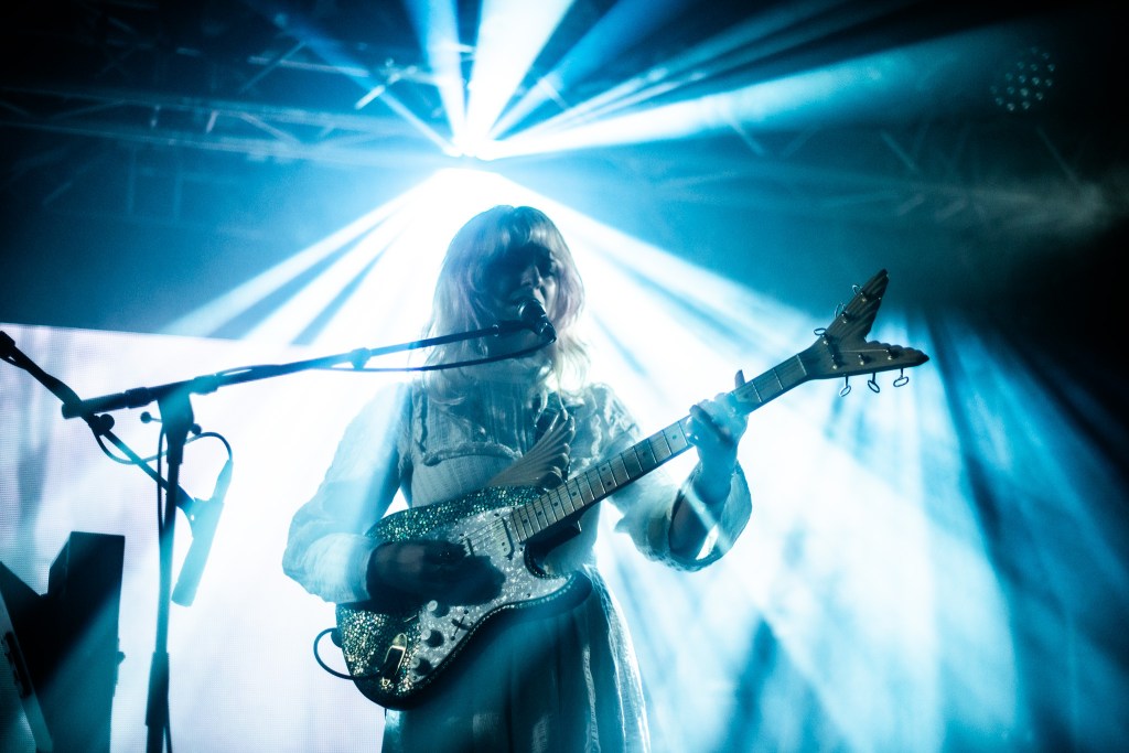 A musician performing on stage, holding a sparkling guitar with blue and white lighting effects in the background.